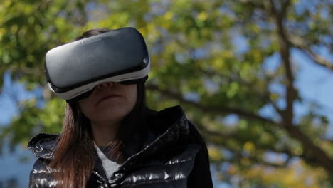 Serious-young-dark-haired-woman-in-warm-black-waistcoat-wearing-virtual-reality-glasses-in-early-autumn-park